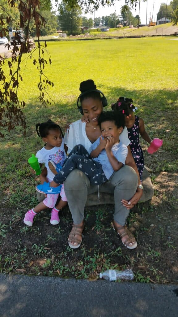 Woman sitting on a stone with three young children, all smiling and holding colorful cups, outdoors in the shade.