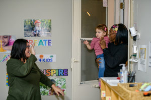 A woman waves goodbye to a child and caregiver at a daycare entrance.