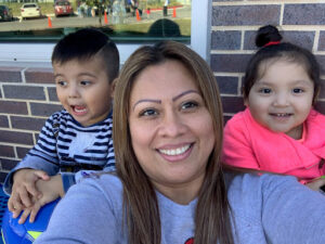 Woman taking a selfie with two smiling young children in front of a brick wall.