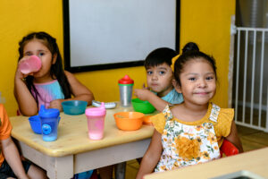 Three young children sit at a small table, eating snacks and drinking from cups, with a yellow wall behind them.