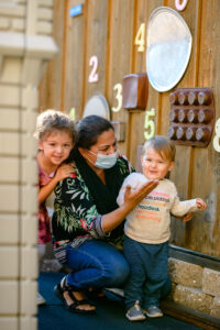 Woman with a mask kneels beside two smiling children in front of a wall decorated with colorful numbers.
