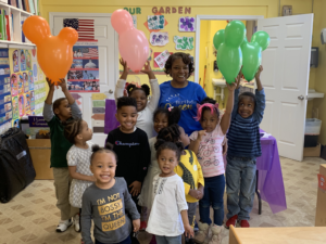 A group of smiling children and an adult hold colorful balloons in a classroom decorated for a birthday party.